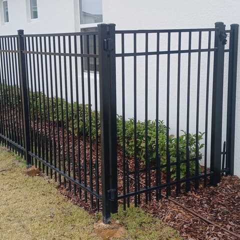 Black aluminium picket fence and gate beside a house, over a mulched bed