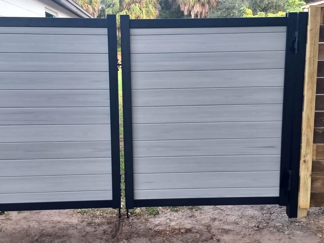 Un portón doble de entrada en tabla compuesta gris sobre marco de acero negro, colgado a escuadra entre sus postes